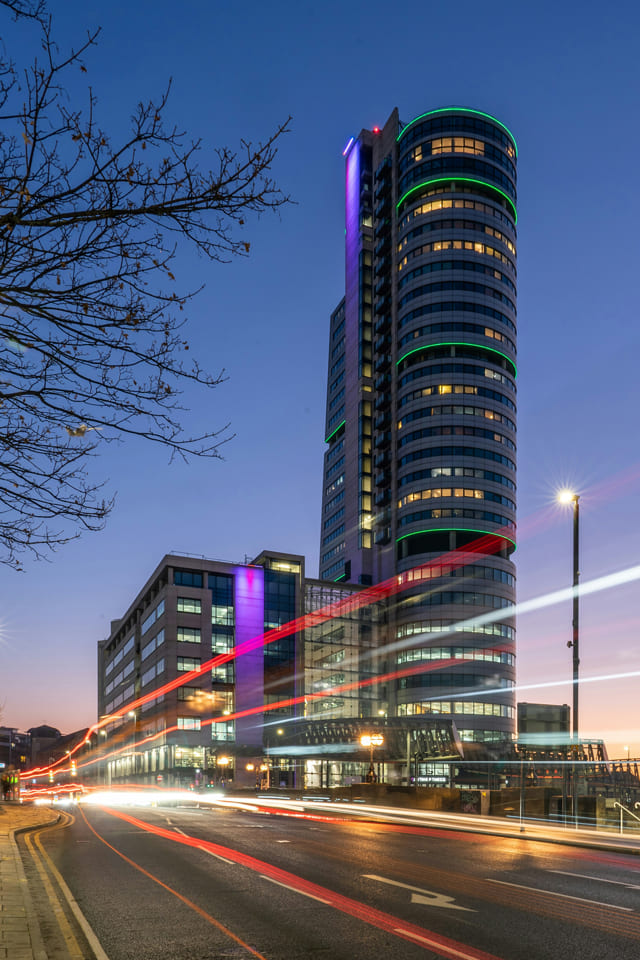 Leeds cityscape at twilight featuring a tall modern glass tower with colourful lights, busy road below with long light trails from passing traffic.