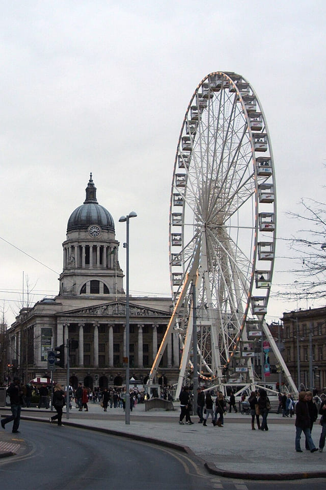Large Ferris wheel beside a historic domed building and busy pedestrian square in Nottingham city centre on an overcast day.