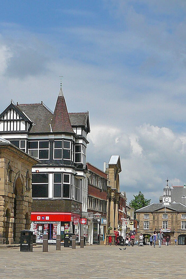 Pontefract street with mixed historic and modern buildings, flower displays, parked bicycles, and people walking under a cloudy sky.