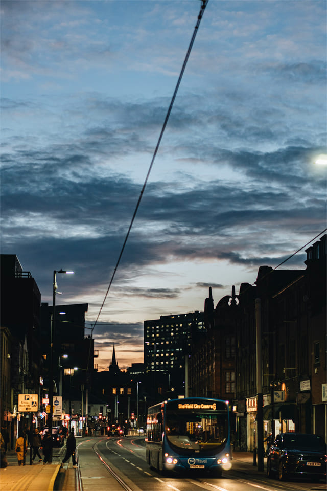 Evening city street with a bus heading to Sheffield City Centre, tram tracks, and pedestrians under a dramatic blue and orange sunset sky.
