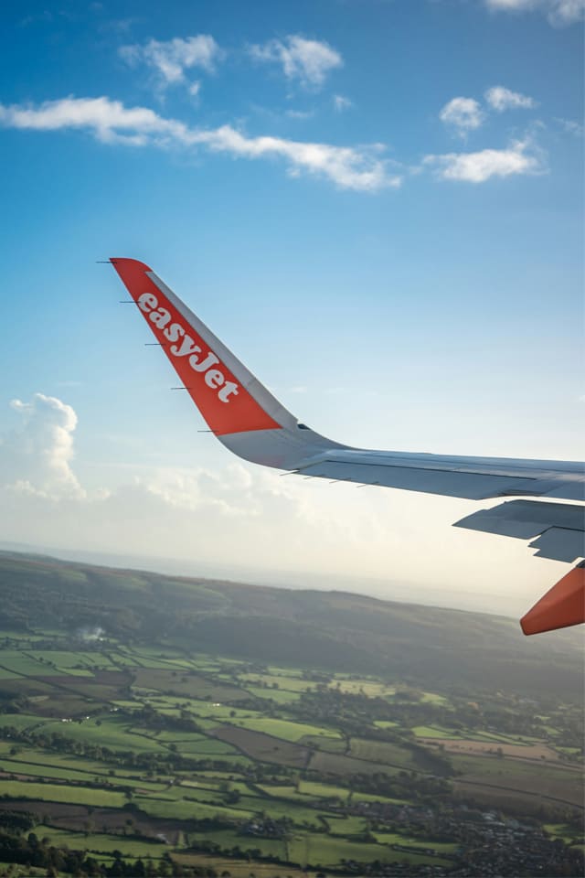 View from an airplane window showing an easyJet wing above a patchwork of green fields under a bright blue sky with scattered clouds.